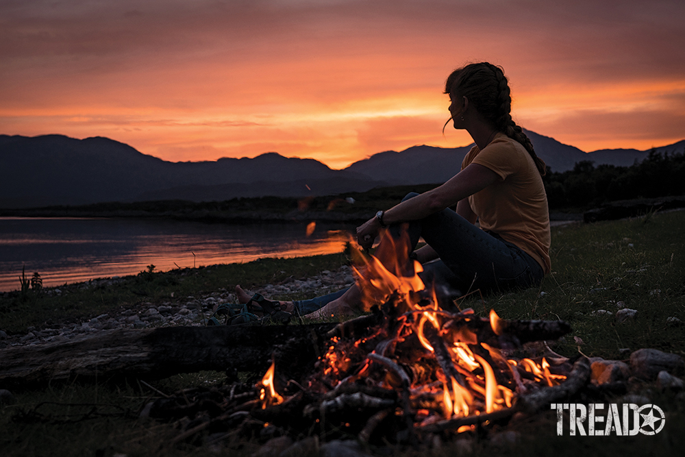 Pink and orange skies give way to a hilly backdrop while a woman sits next to a campfire on a beach.