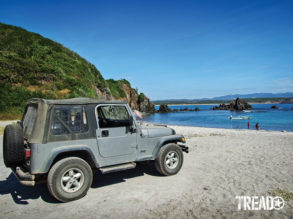 Silver two-door soft-top Jeep sitting on beach next to lush hillside.