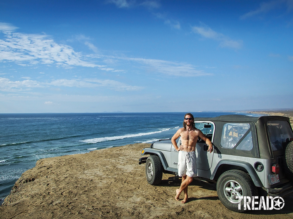 Shirtless man with white shorts standing next to silver Jeep next to ocean on beach.